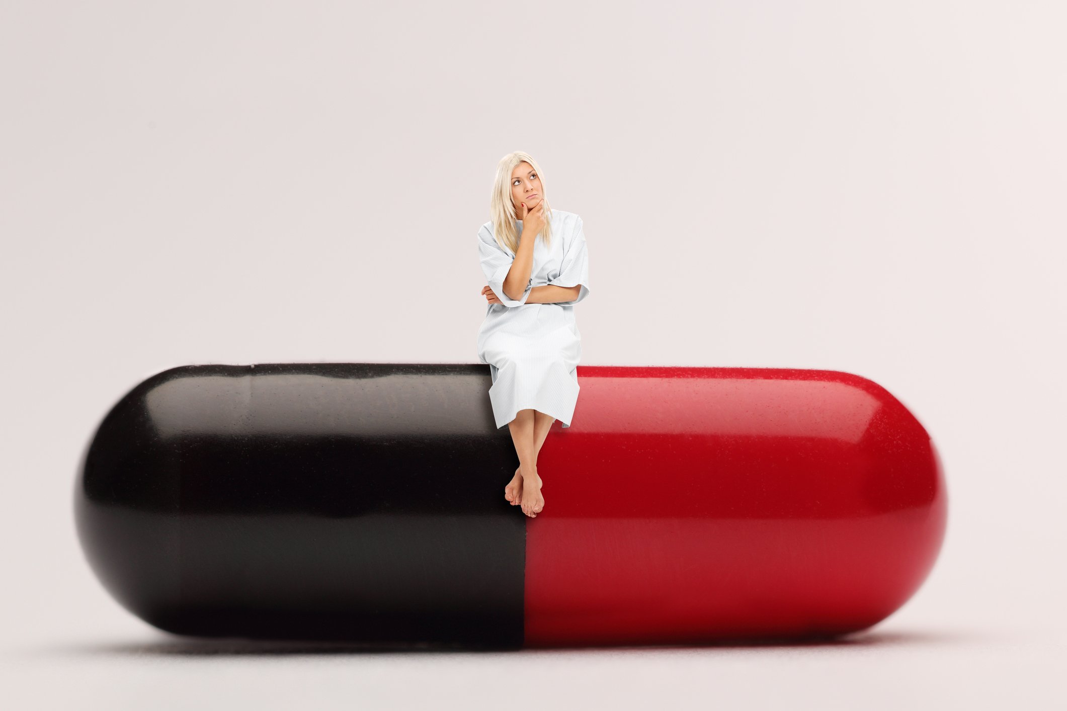 Patient in hospital gown sitting on a giant black-and-red medicine capsule.