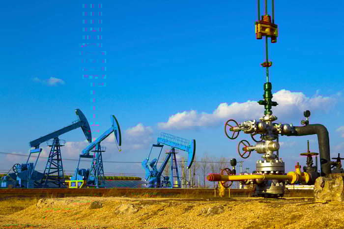 A close-up of a natural gas wellhead with three oil pumps in the background.