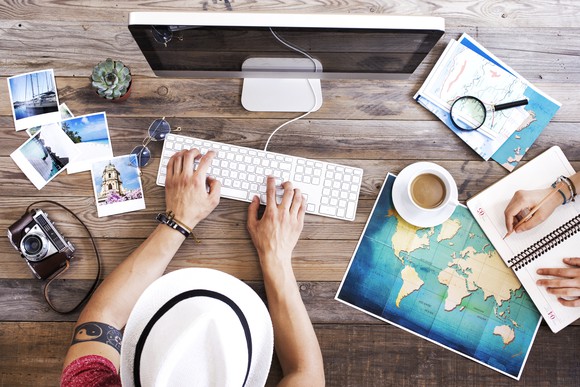 Man in hat at computer surrounded by travel items, including a map, pictures, camera, and notebook.