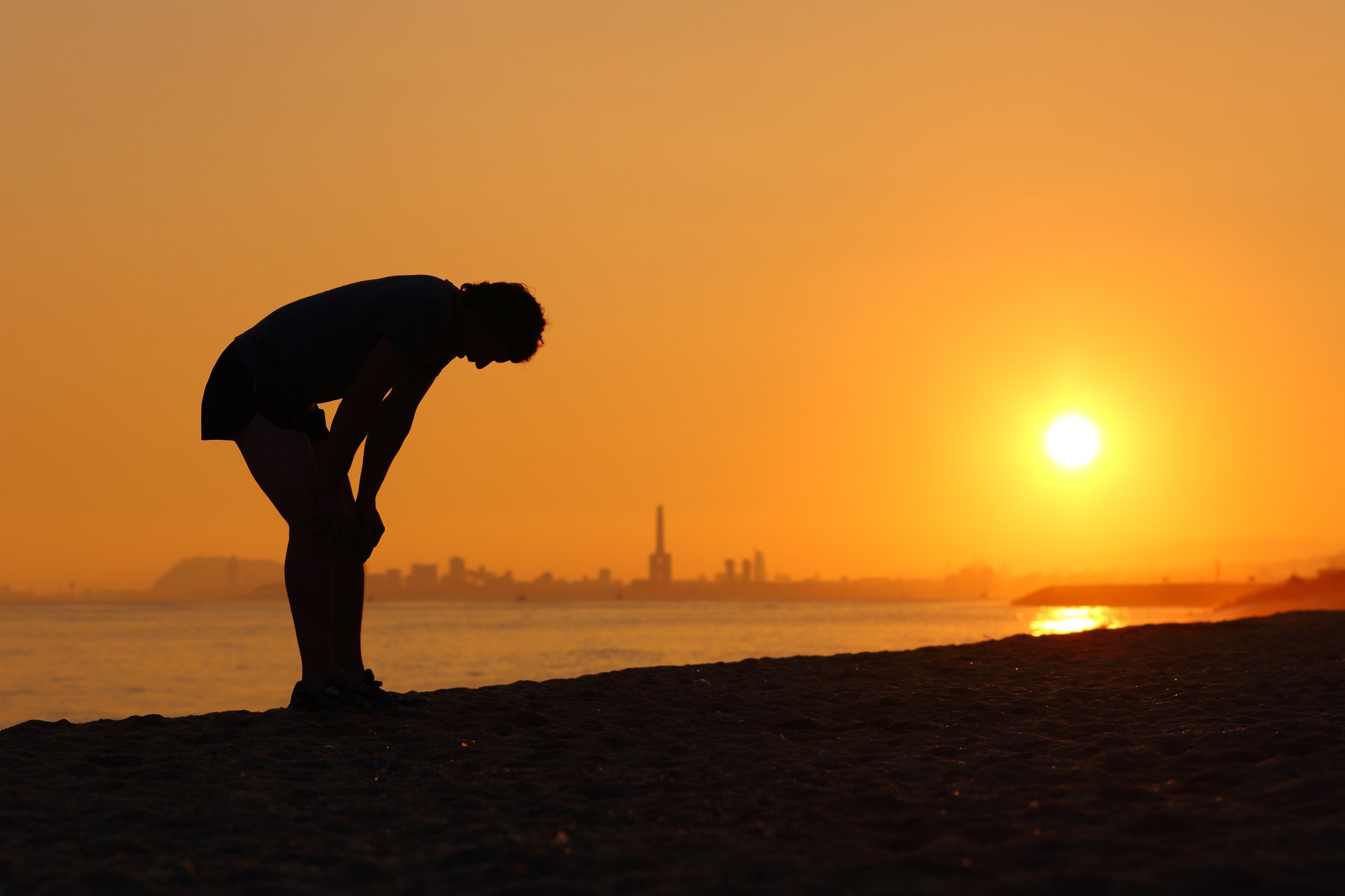 Silhouette of a tired sportsman resting with his hands on his knees in front of a golden Spanish sunset.