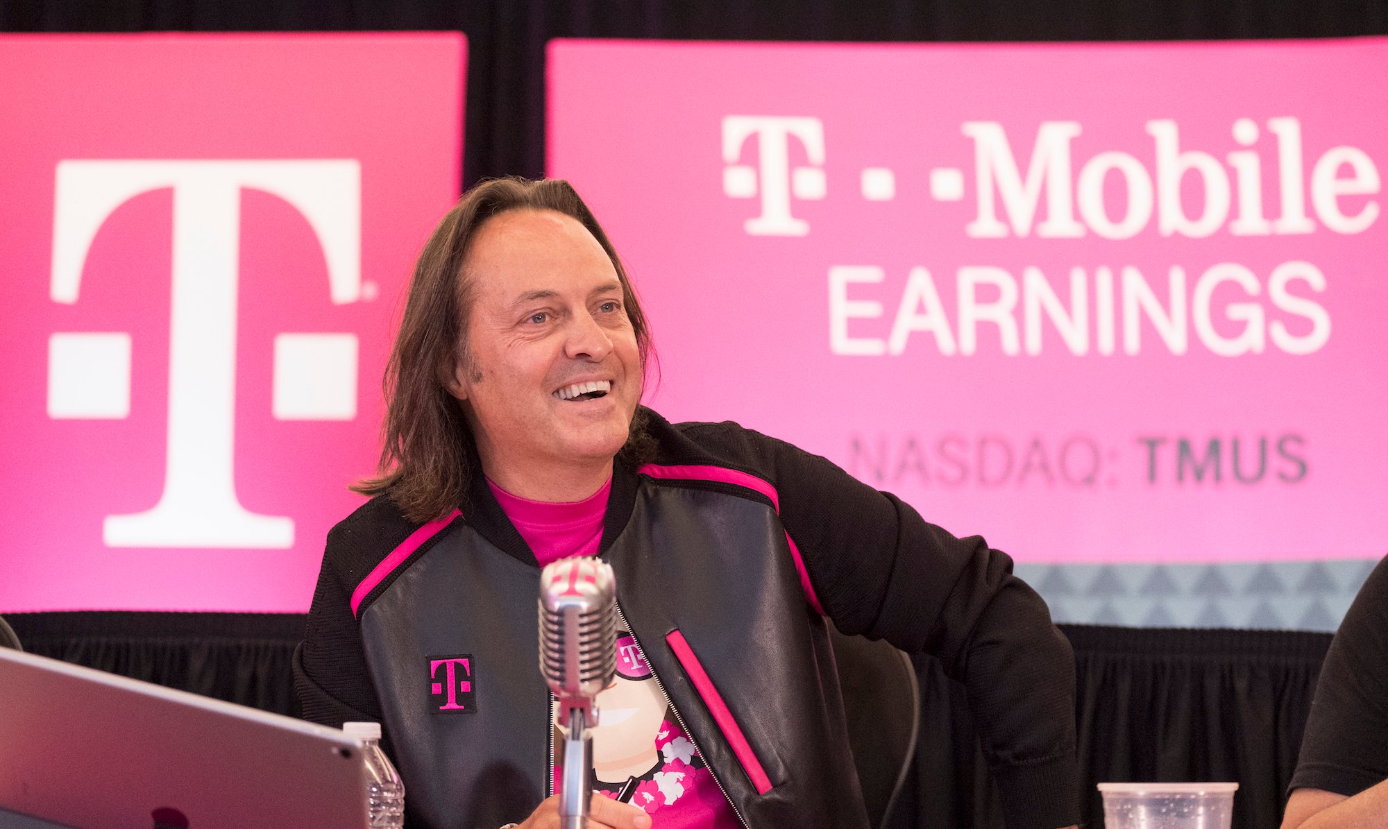 T-Mobile CEO John Legere is at a T-Mobile earnings event sitting behind a microphone smiling.