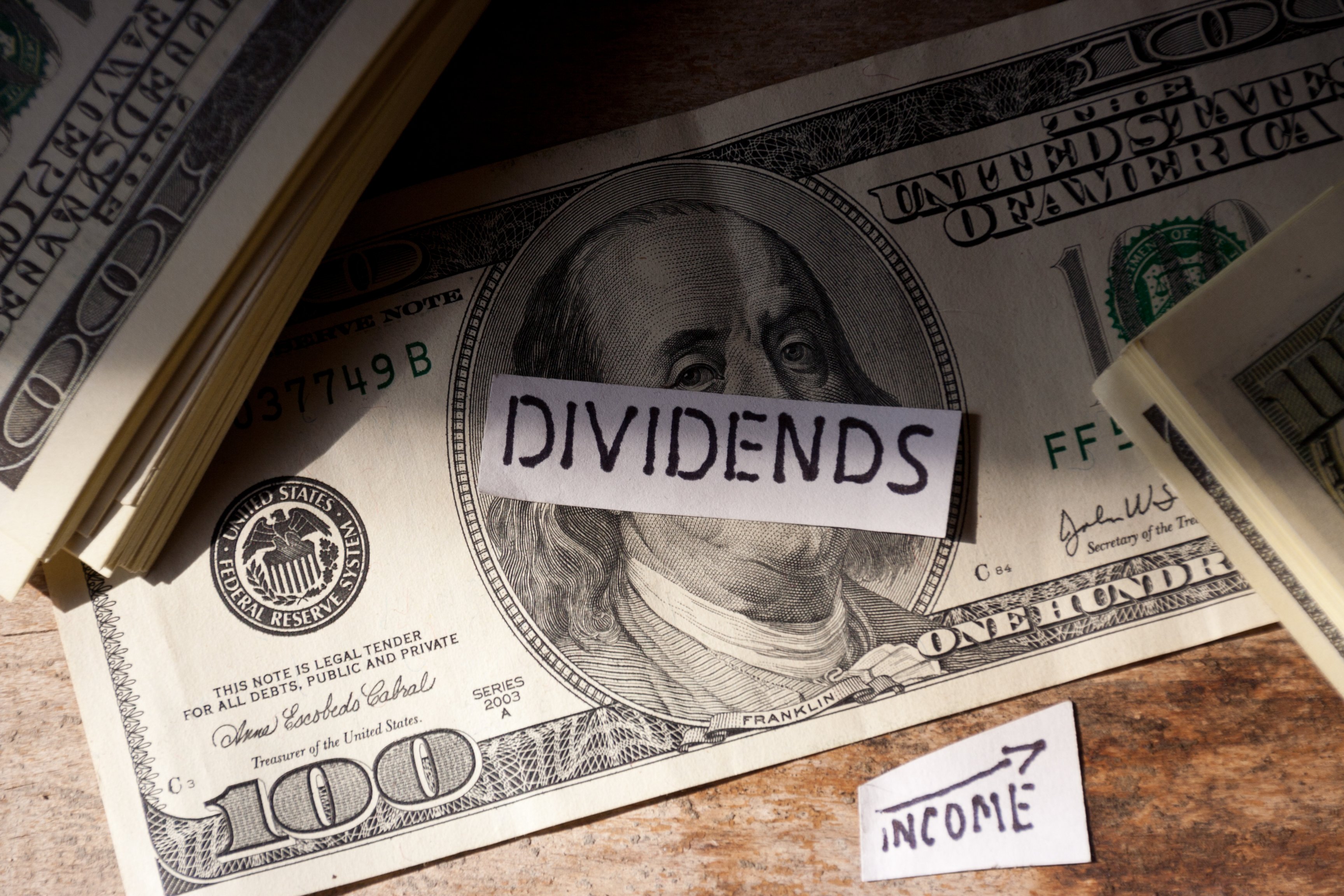 $100 bill with dividends tab on top of it next to two stacks of $100 bills and a small piece of paper with income and a line going up written on it