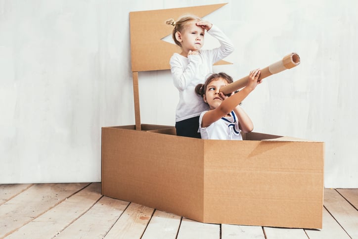 Two children playing pretend in a boat made out of cardboard.
