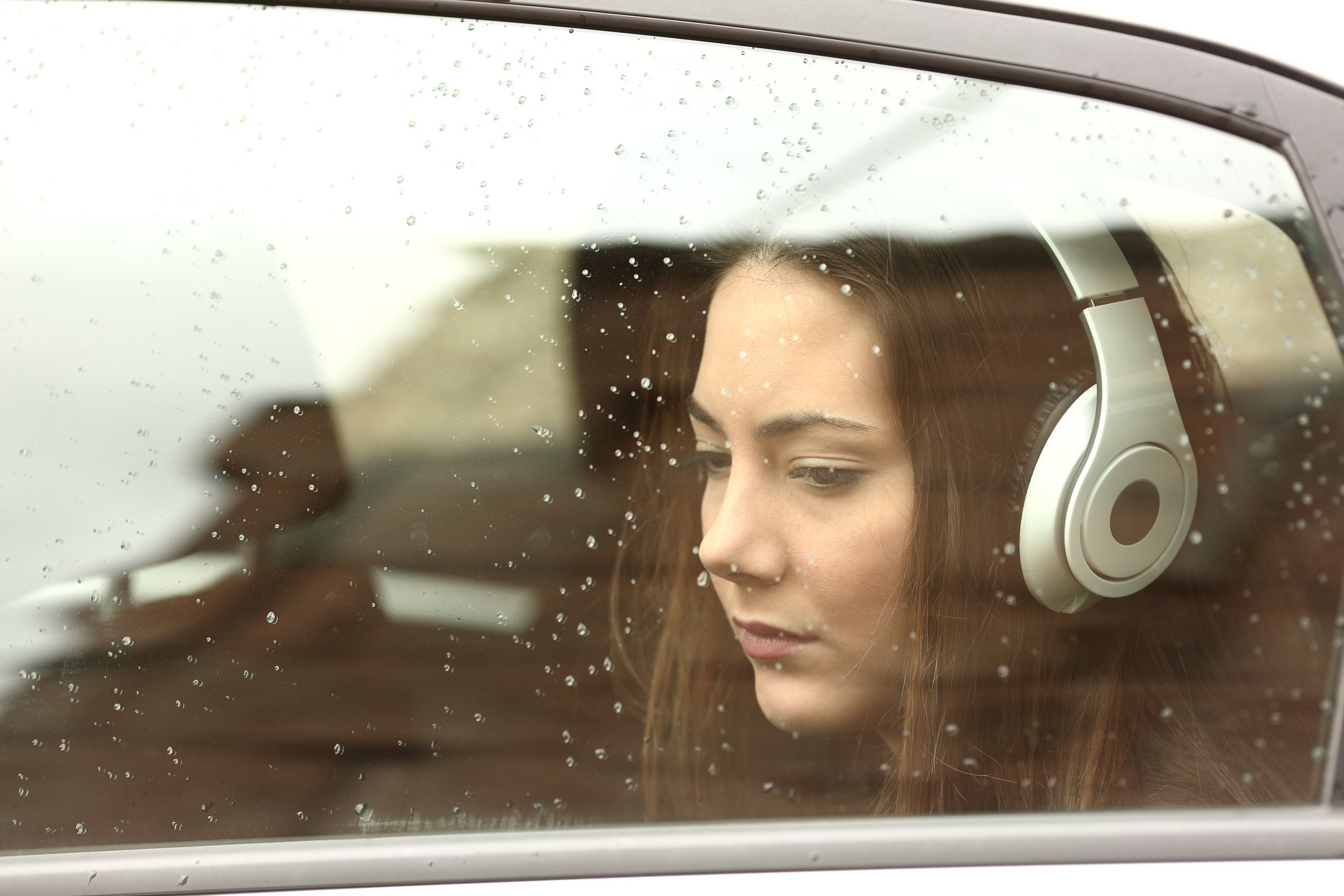 Young woman wearing headphones in the backseat of a rain-covered car.