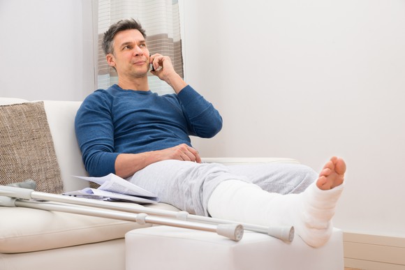 Man with broken leg in cast sitting on couch talking on phone.