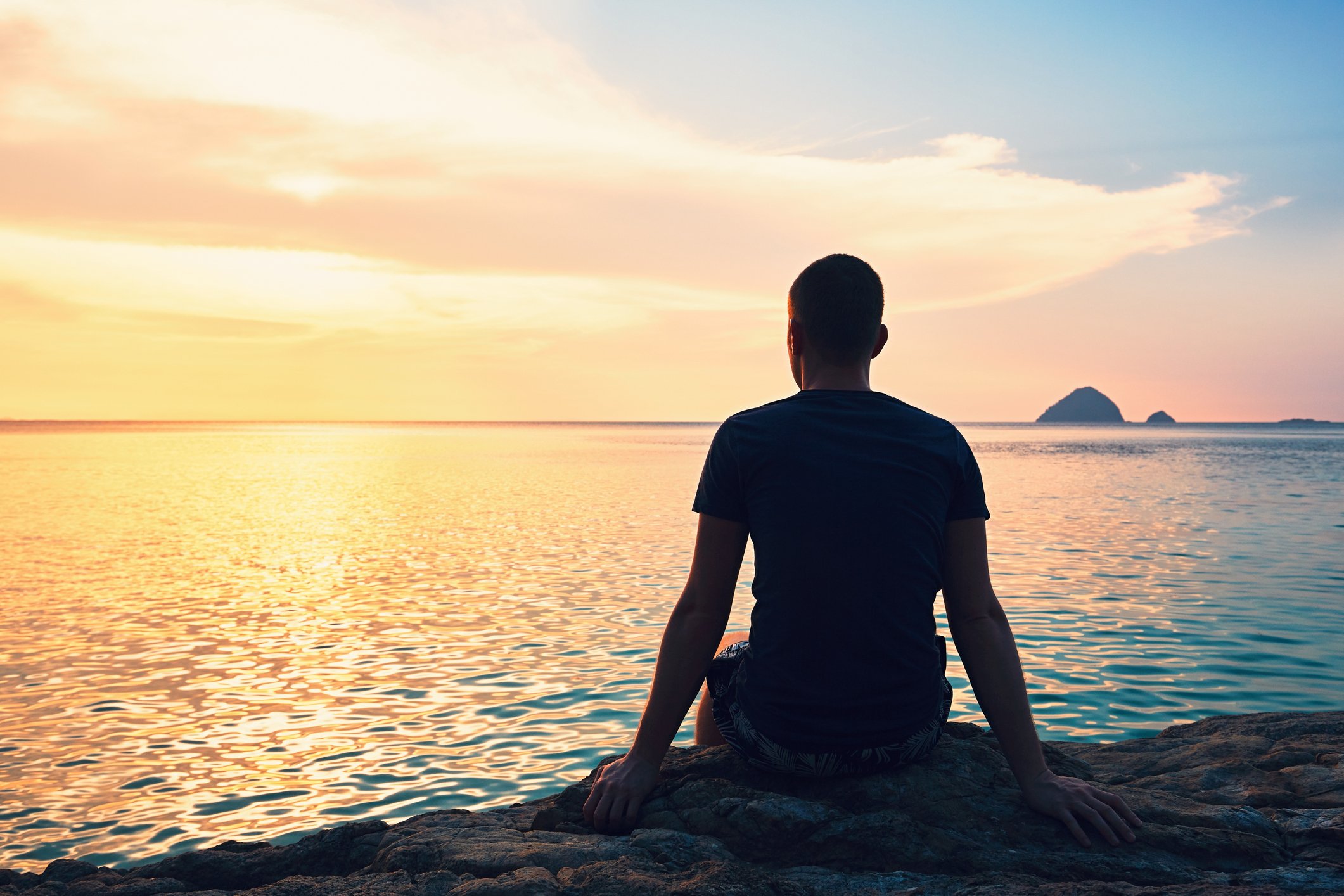 Man sitting facing the water at sunset