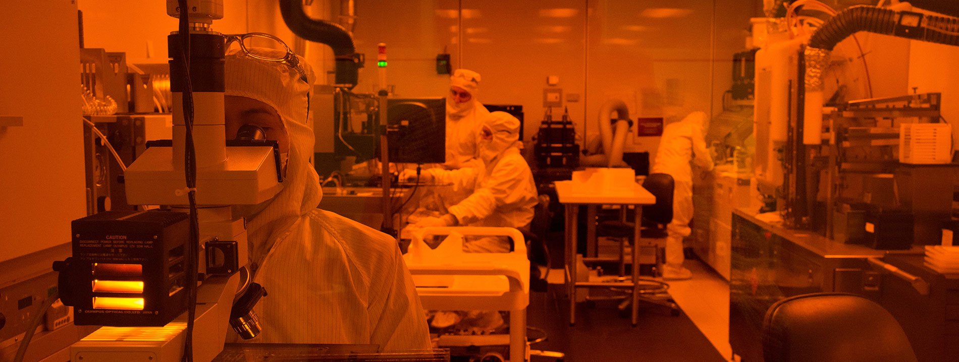 Lab in orange light with people wearing masks and sterile clothing working on various types of equipment.