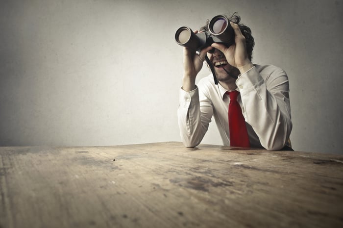 A businessman sitting at a table looking through large binoculars.