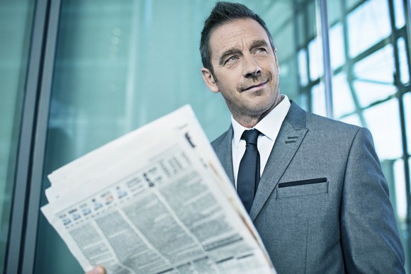 A smirking man in a suit reading a financial newspaper. 
