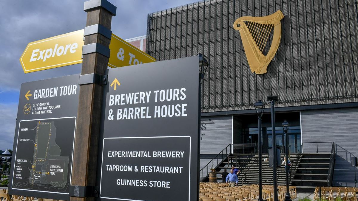 An outside view of Guinness new brewery. Guinness' harp-shaped logo is displayed on top of the building, and signs with directions are displayed in the foreground.