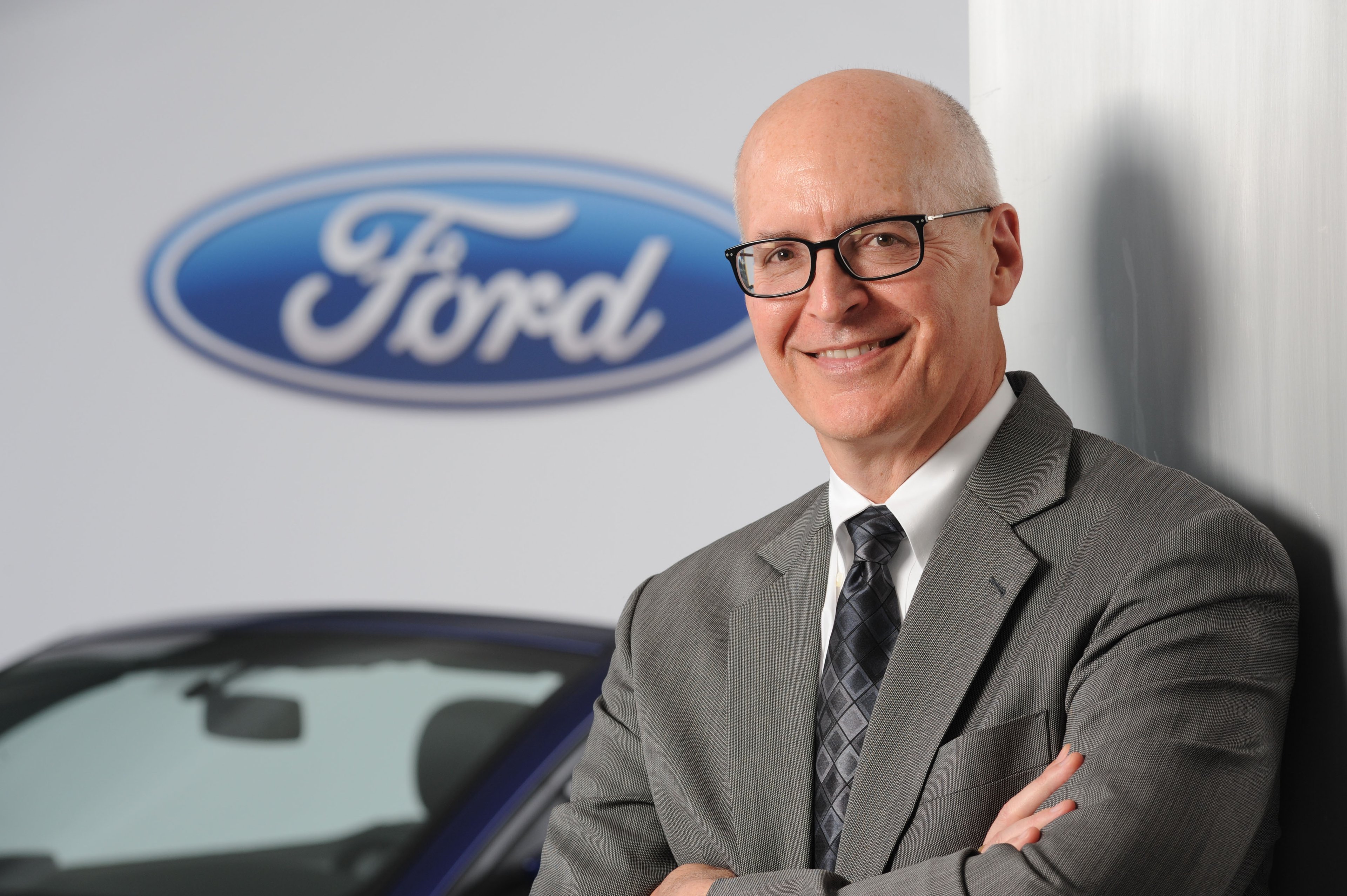 Ford CFO Bob Shanks is shown standing in front of a Ford Mustang and a white backdrop with Ford's logo.