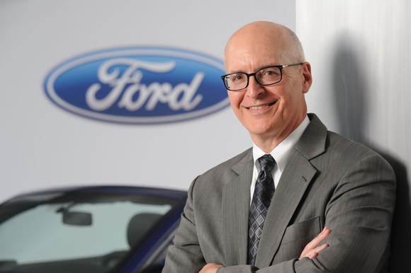 Ford CFO Bob Shanks is shown standing in front of a Ford Mustang and a white backdrop with Ford's logo.