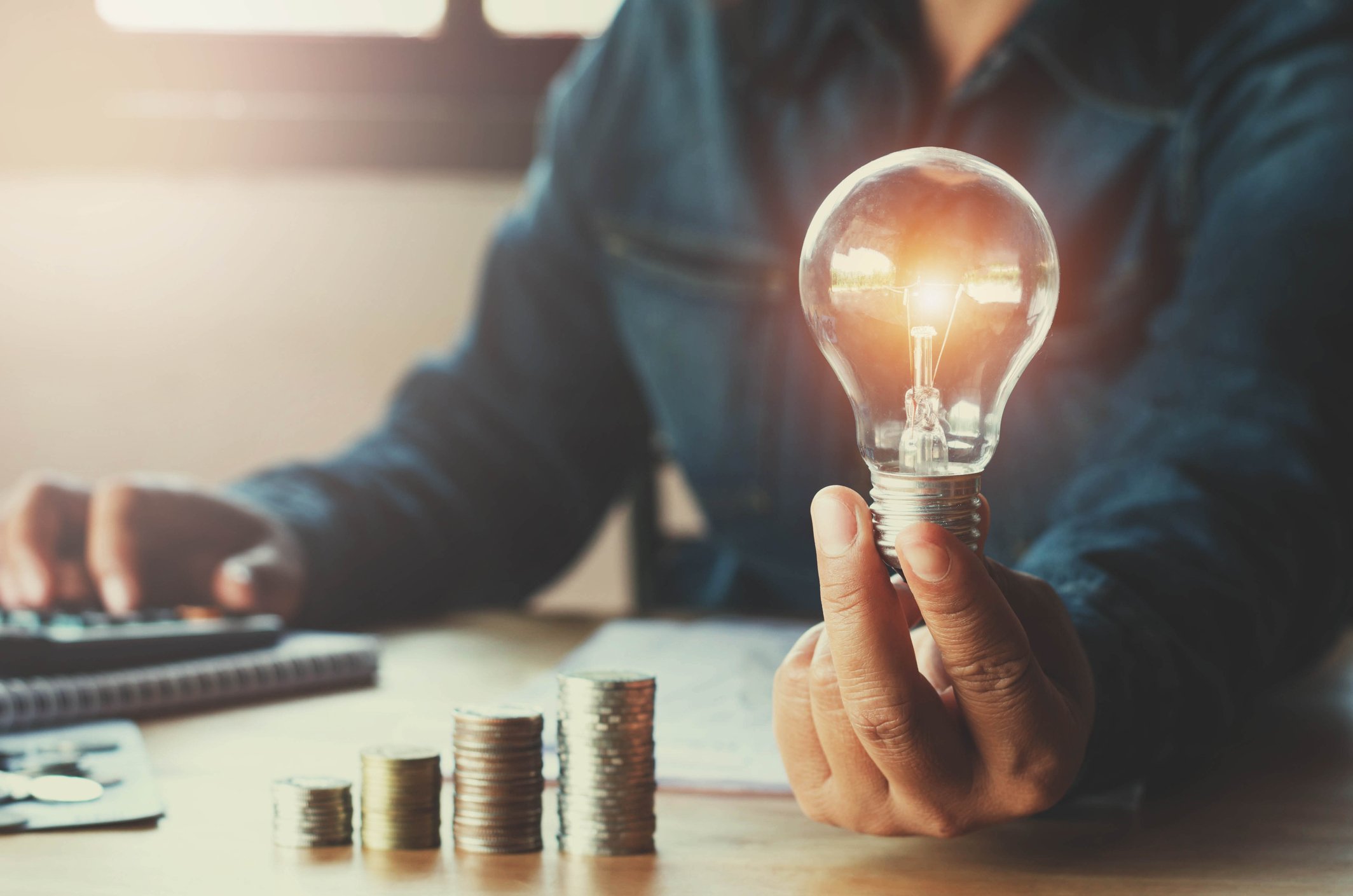 A man using a calculator while holding a light bulb in front of an ascending stack of coins