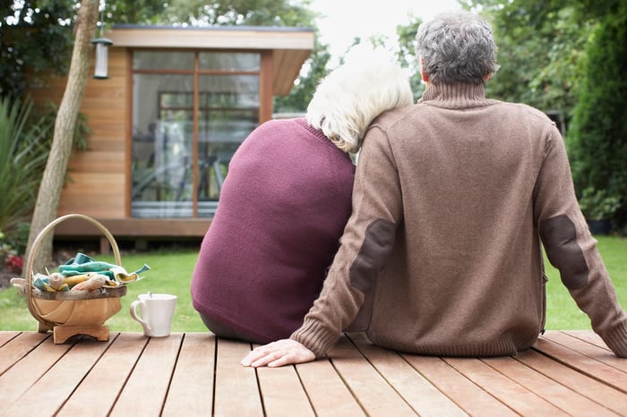 Older man and woman sitting on a deck. Woman rests her head on the man's shoulder.