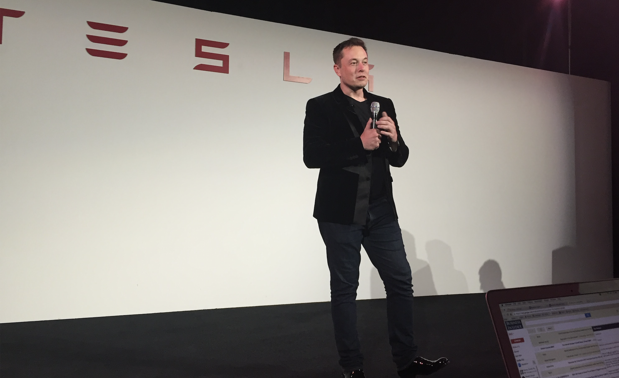 Elon Musk is standing on a stage holding a microphone and talking to an audience.