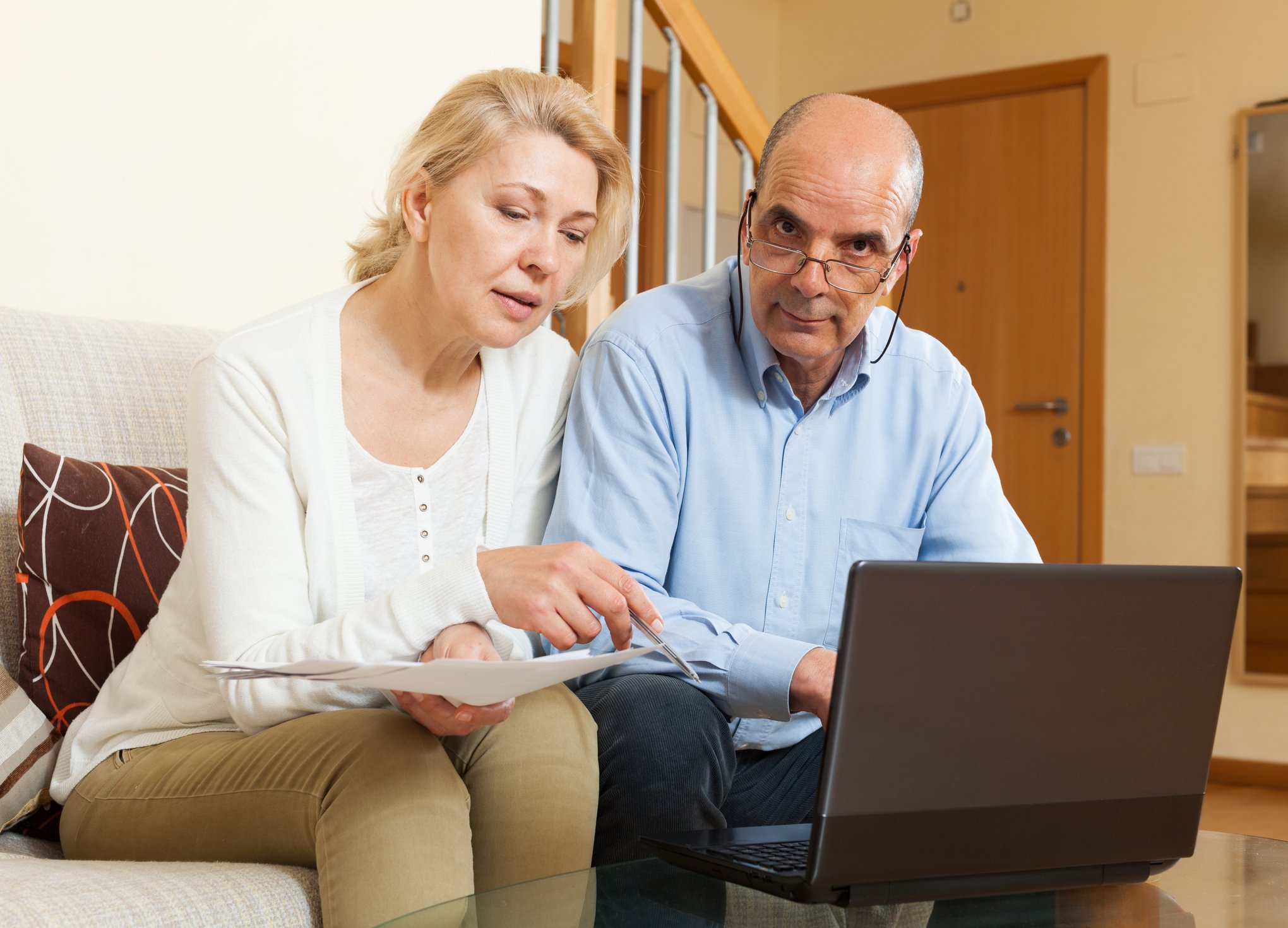 A mature couple examining their finances on a laptop, with the husband visibly irritated. 