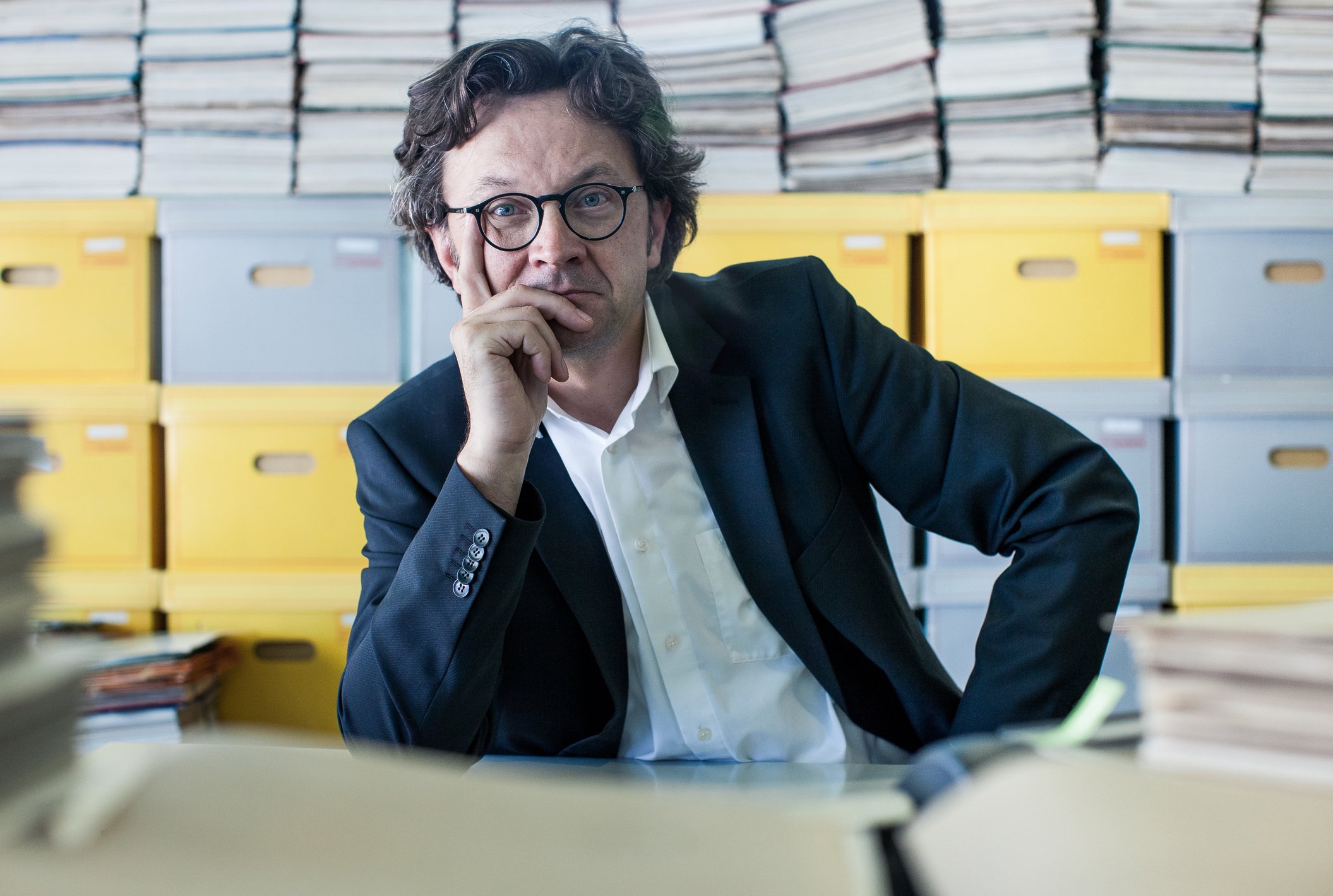 A man in his 40s in deep thought, with his head resting on his right hand and numerous file cabinets behind him.