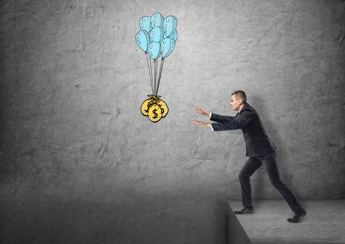 A businessman reaching for bags of money hanging in the air just over a cliff.