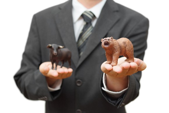 Man in a business suit holding a bear figurine in one hand a bull figurine in the other.