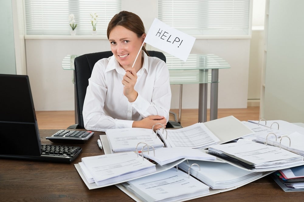 Professional woman sitting at her messy desk while holding up a small sign with the word help on it.
