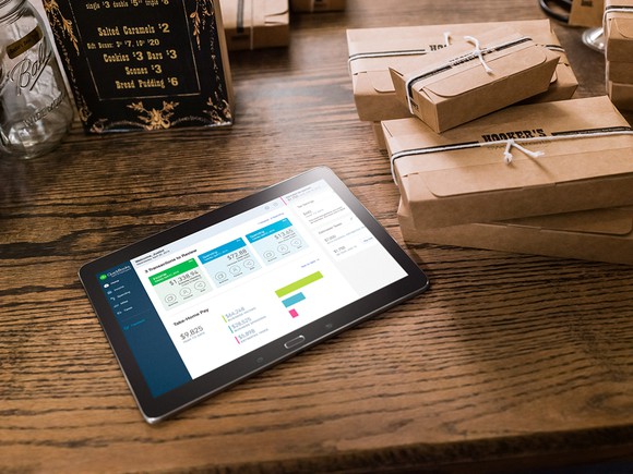 QuickBooks Online on a tablet that's sitting atop a wooden table and alongside boxes wrapped with brown paper.