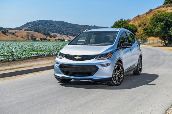 A silver 2018 Chevy Bolt EV drives on a road with mountains in the background