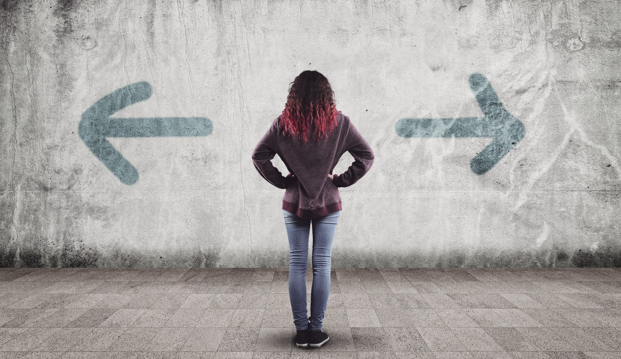 A woman faces a wall with her hands on her hips, as she stands between arrows pointing in opposite directions.