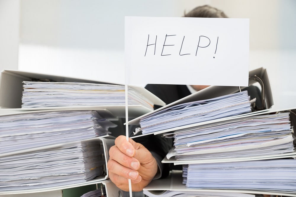Man at desk buried in paperwork holds up small sign with Help! written on it.