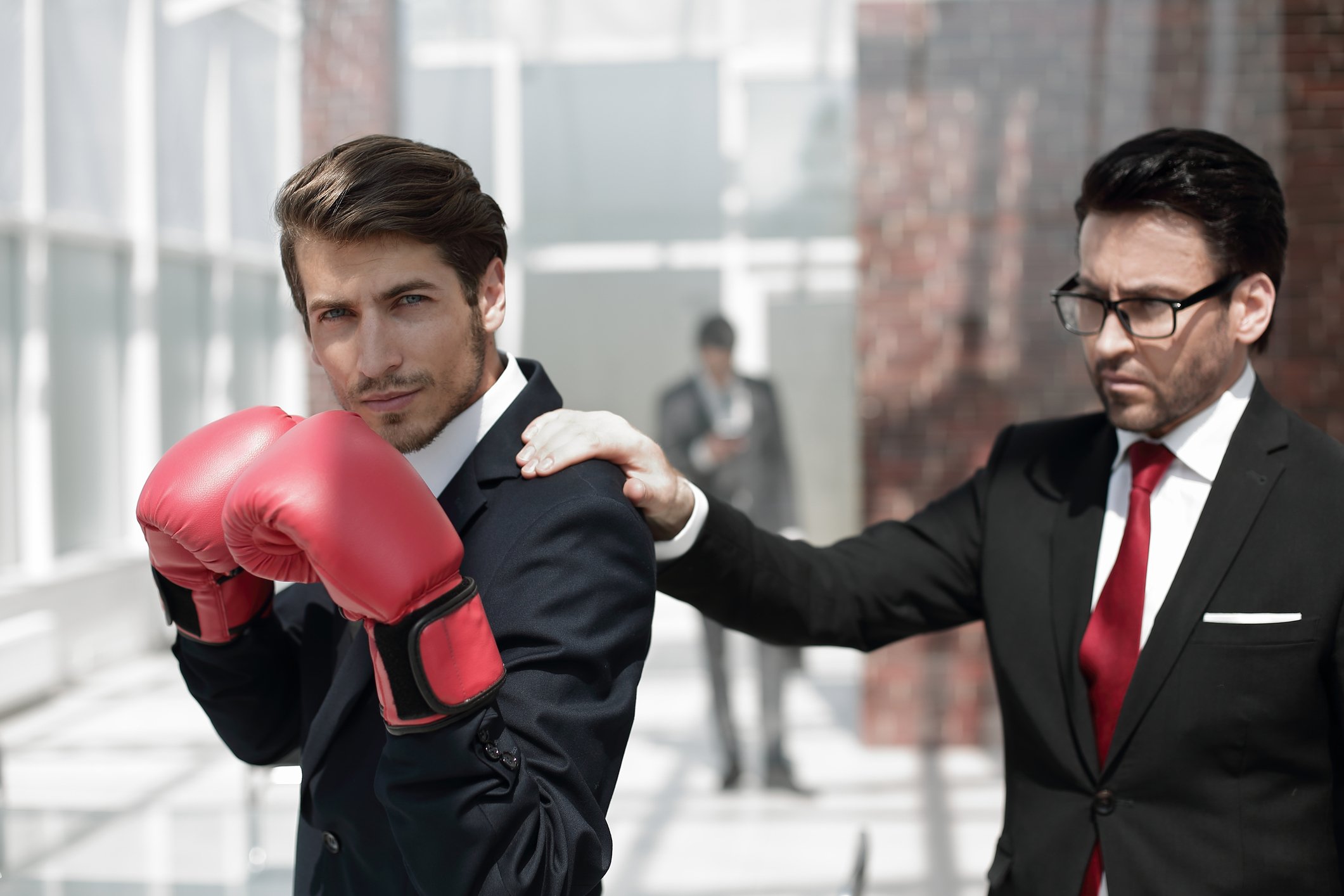 One businessman wearing boxing gloves while another puts a calming hand on the boxer's shoulder.