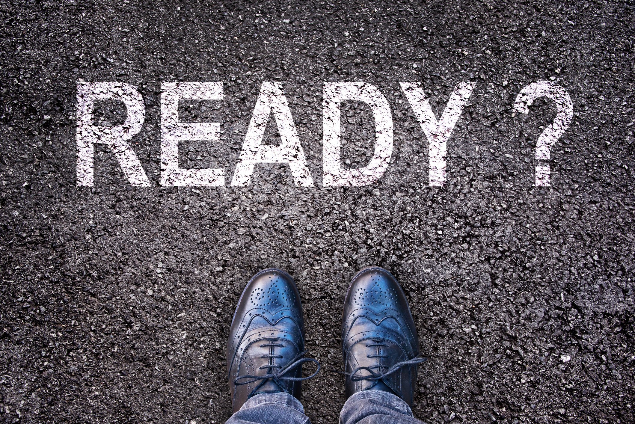 The word ready with a question mark printed on asphalt, with two feet in shoes showing, facing the word
