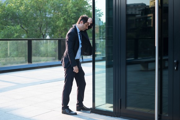 A businessman in a suit rests his head sullenly against a glass wall.