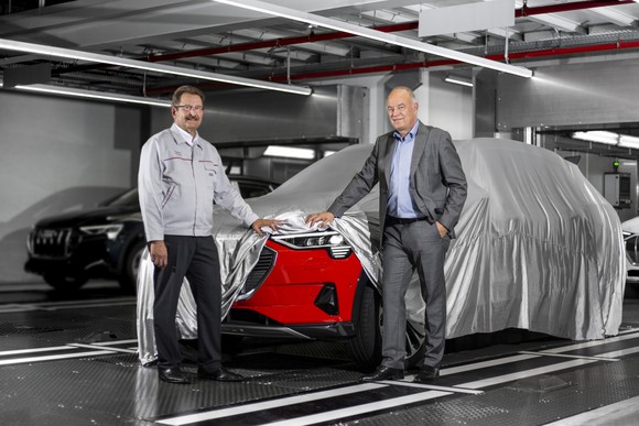 Patrick Danau and Peter Koessler standing at the end of an automotive assembly line, with a vehicle under a show-car-style cover. One corner of the cover is lifted to reveal the front of a red Audi SUV.