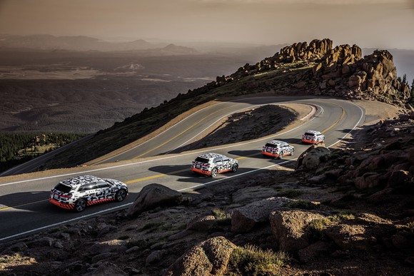 Four Audi e-tron prototypes, in their distinctive black-white-orange camouflage, are shown heading downhill on a winding mountain road.