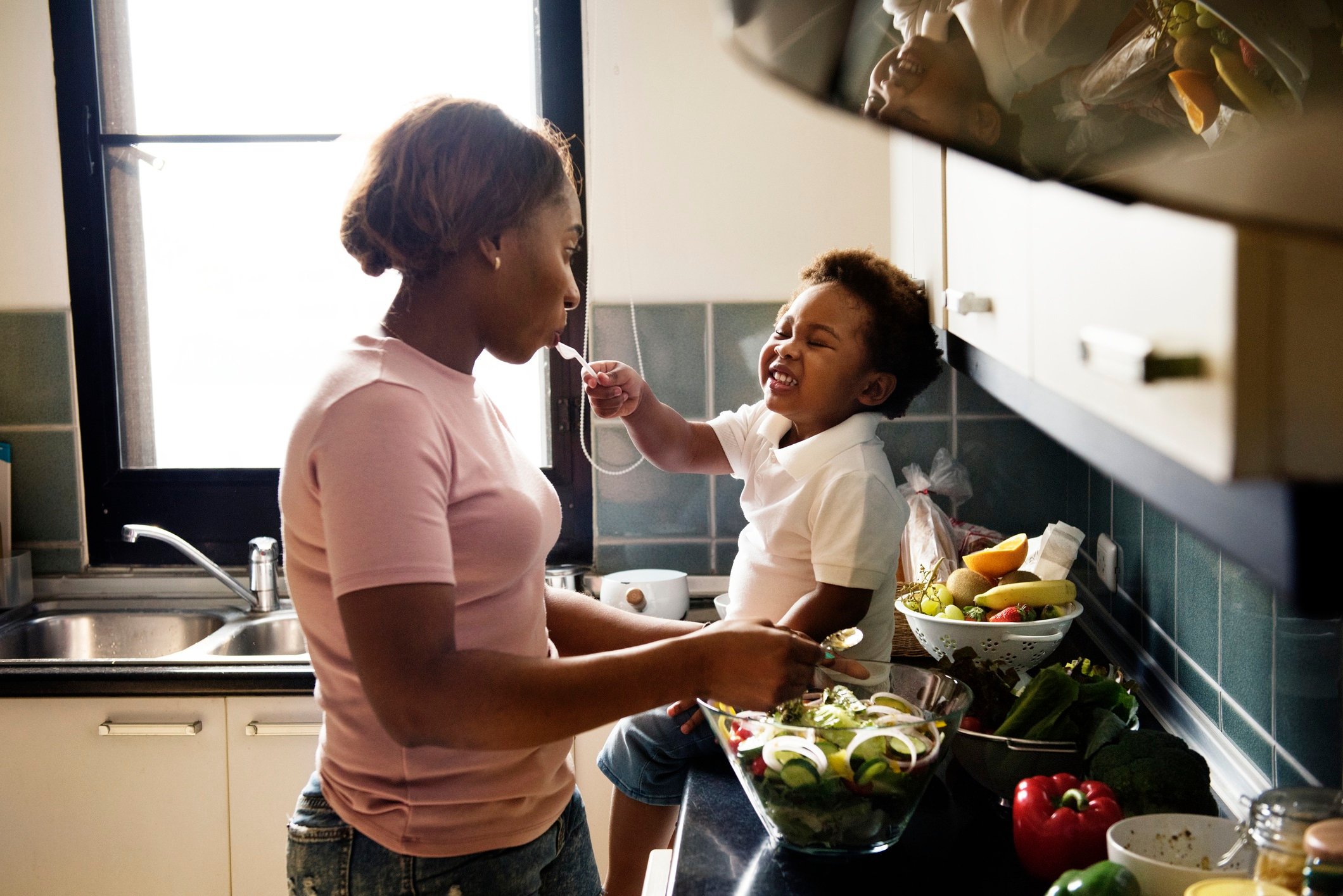 Woman standing near a kitchen counter preparing a fresh salad while a toddler sitting on the counter smiles and puts a spoon in the woman's mouth