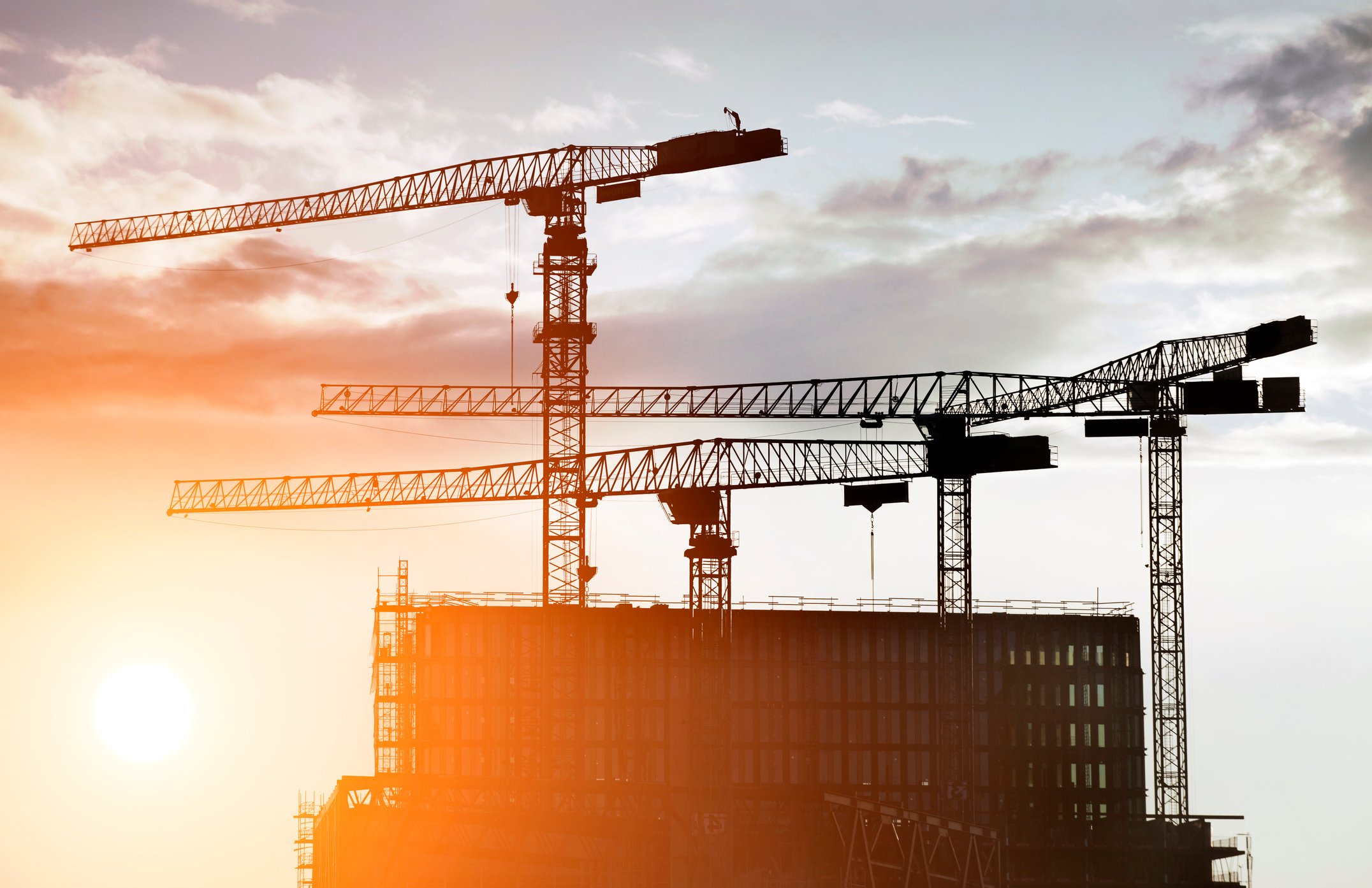 The sun rising behind a building with cranes surrounding it.