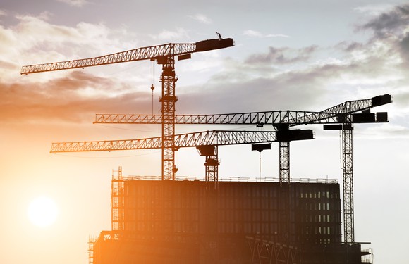 The sun rising behind a building with cranes surrounding it.