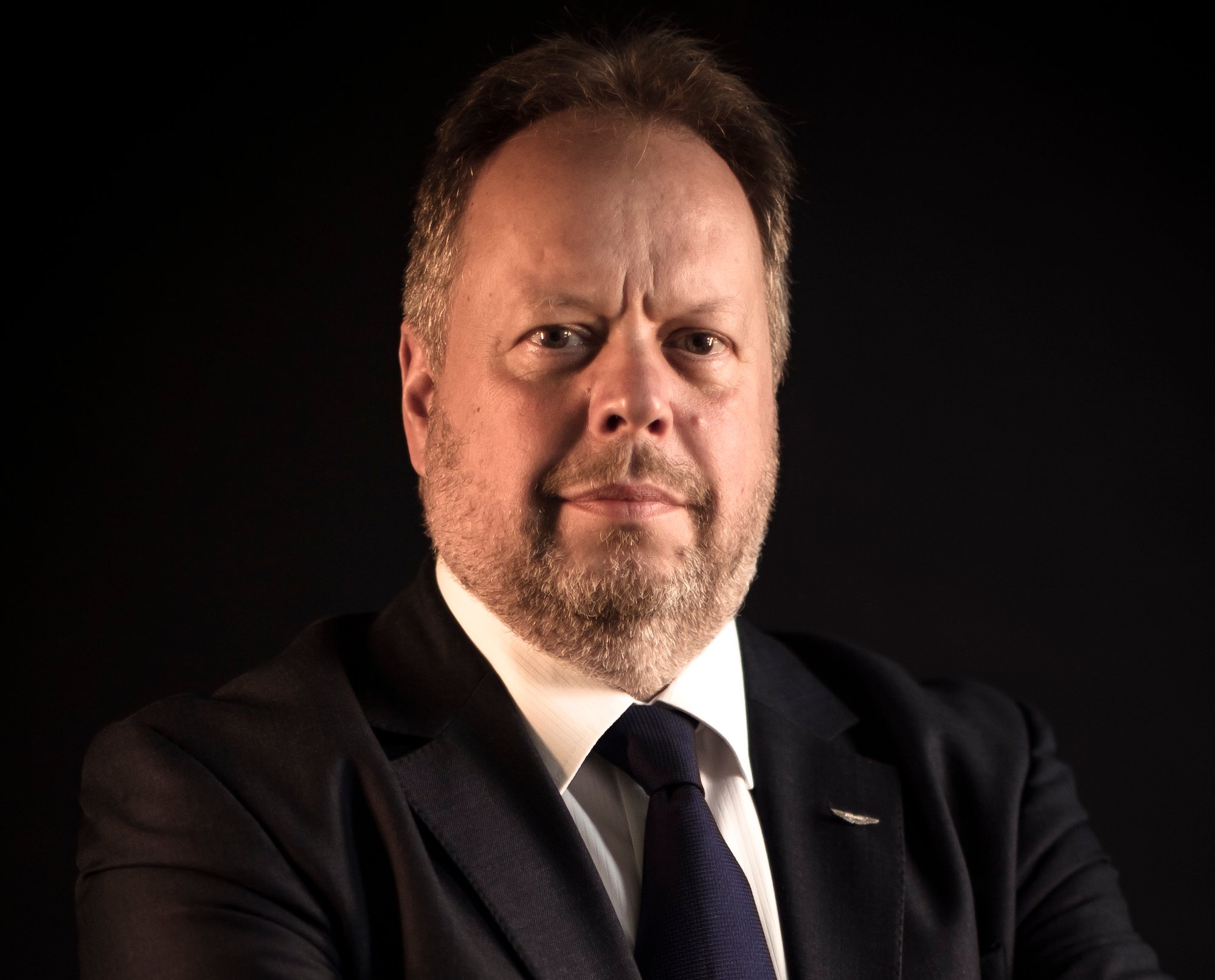 Palmer is shown wearing jacket and tie, seated before a black backdrop.