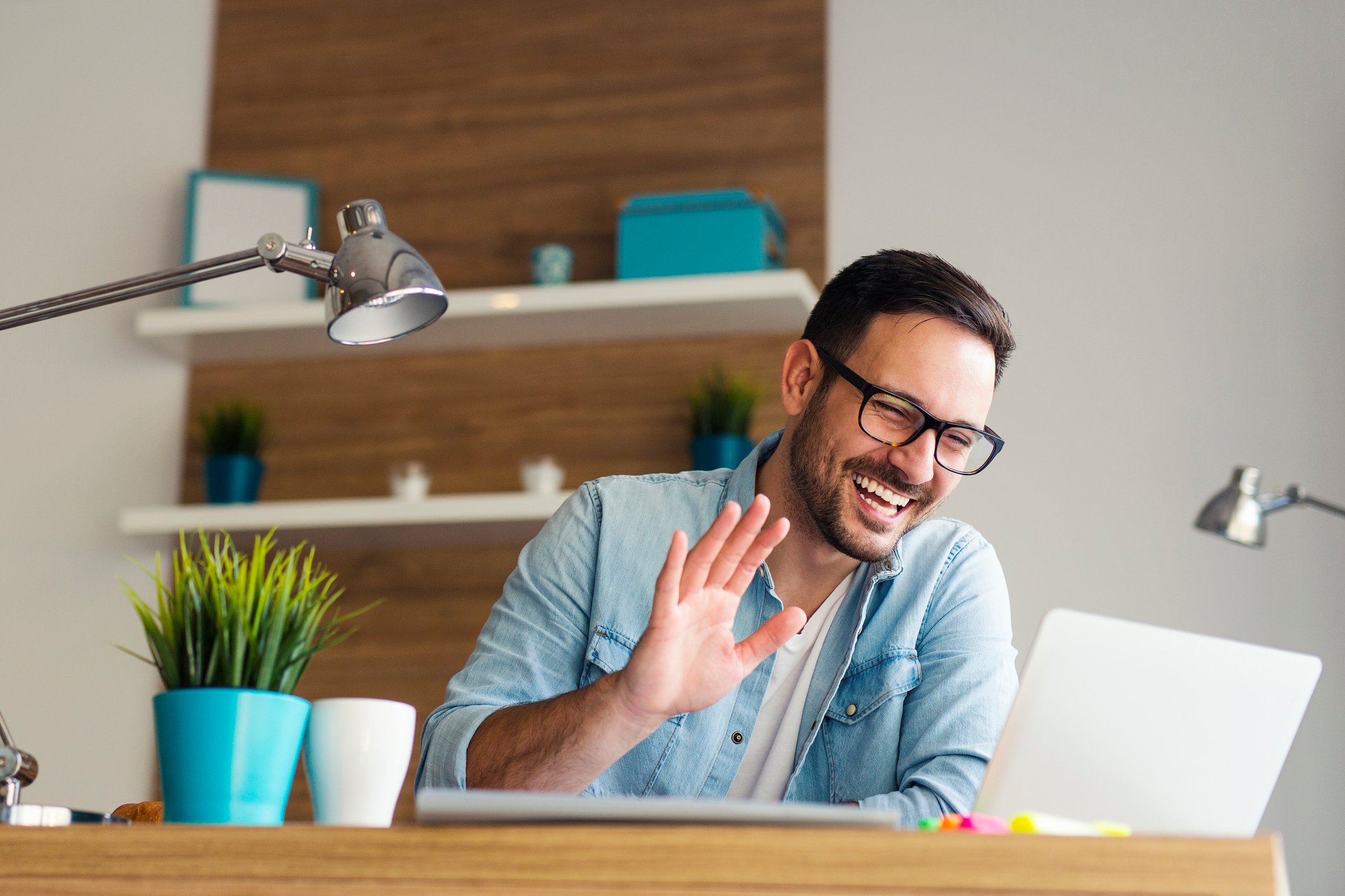 A smiling young man waving at the screen of a laptop computer