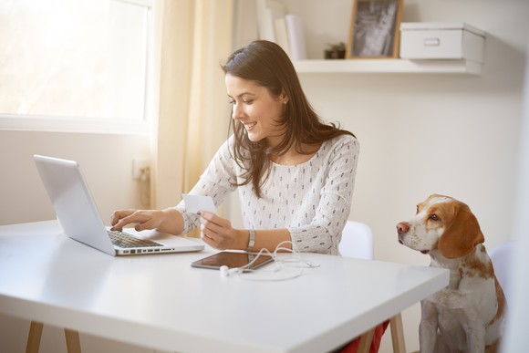 Woman at a desk working at a laptop while a dog looks on.