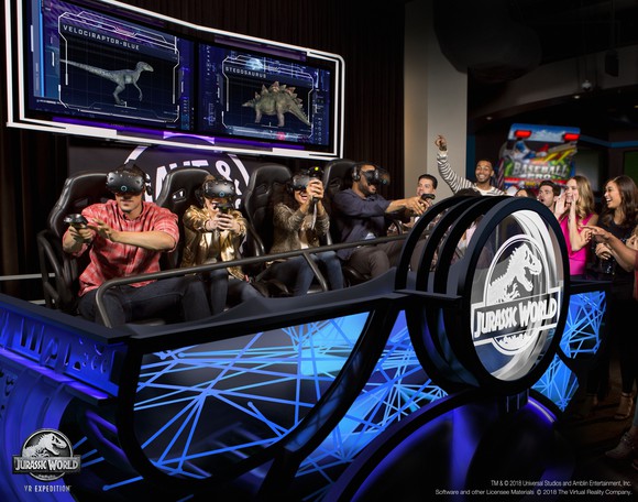 Four people playing Jurassic World VR Expedition. Onlookers cheer the gamers on.