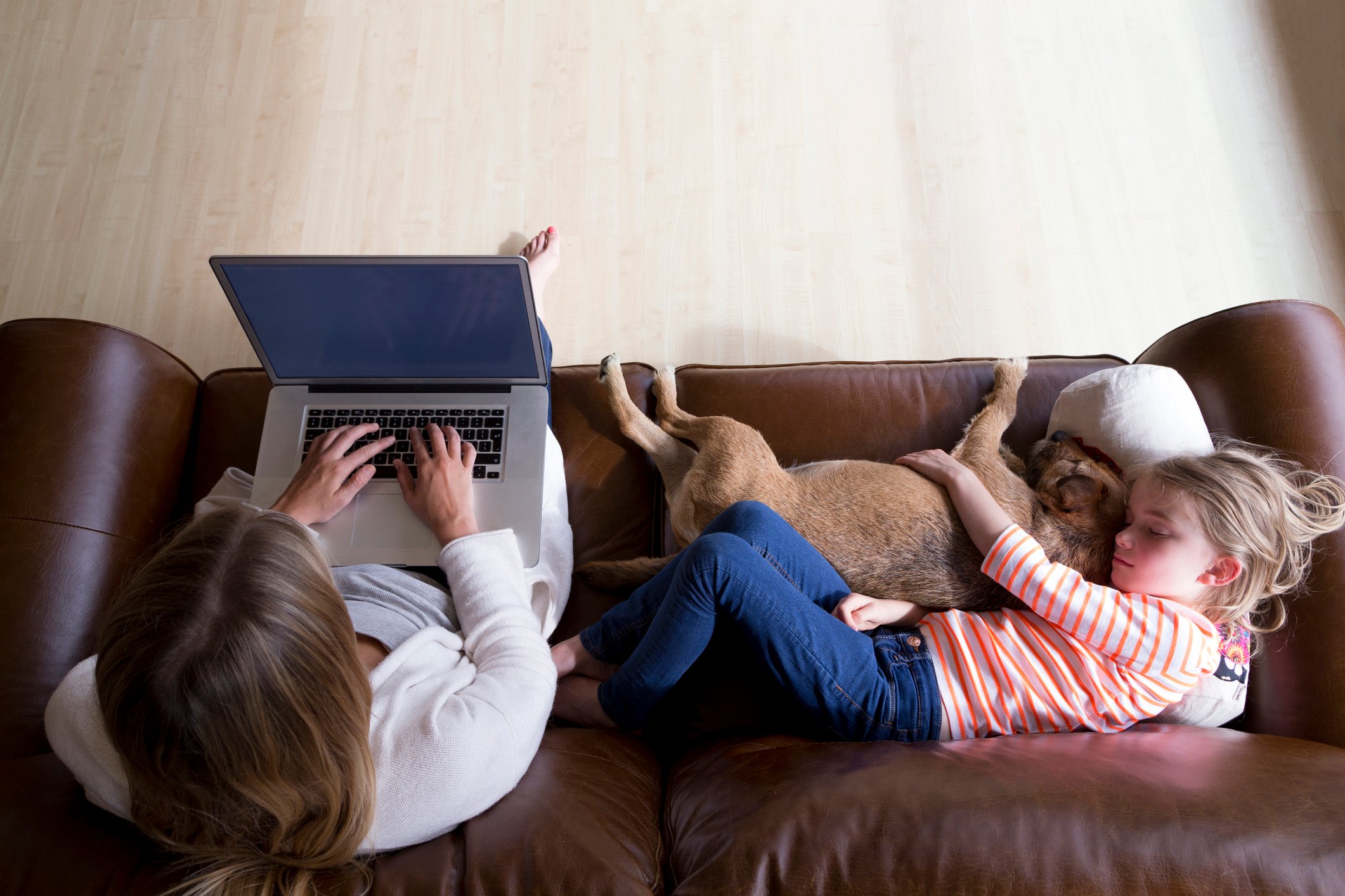 A woman types on a laptop next to a child cuddling a dog.