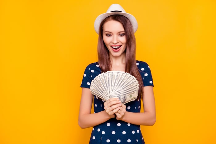 Smiling young girl in a blue dress and hat fanning hundred-dollar bills in front of a lemon-yellow wall.