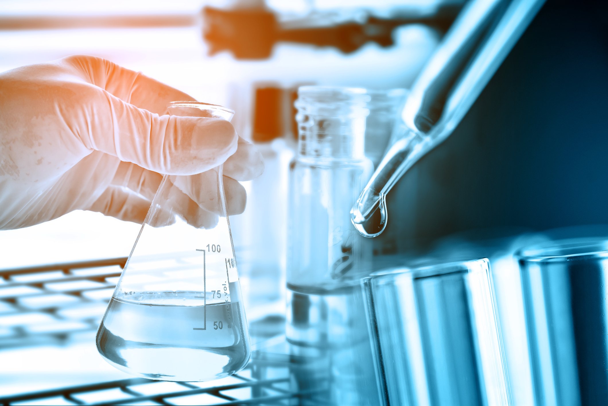 Gloved hand holding flask with dropper in foreground and test tubes and bottles in background.
