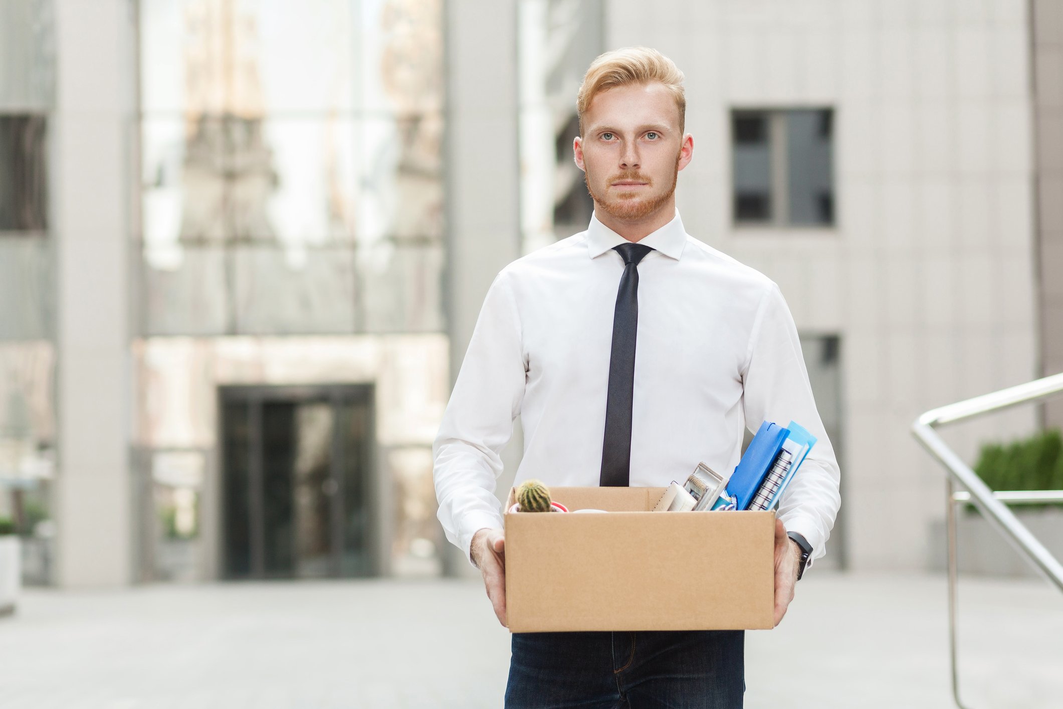 Man in dress shirt and tie carrying cardboard box of office supplies.