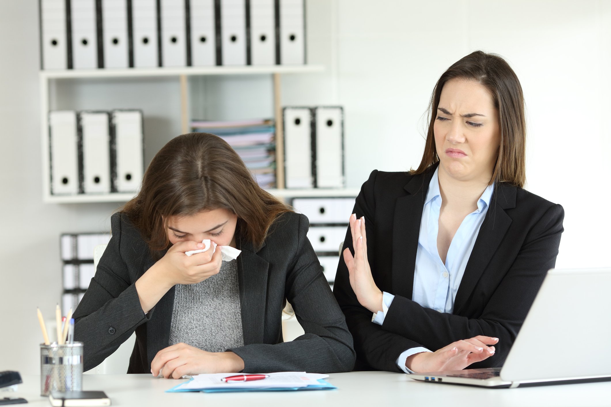 A woman looks on as another sneezes in the workplace