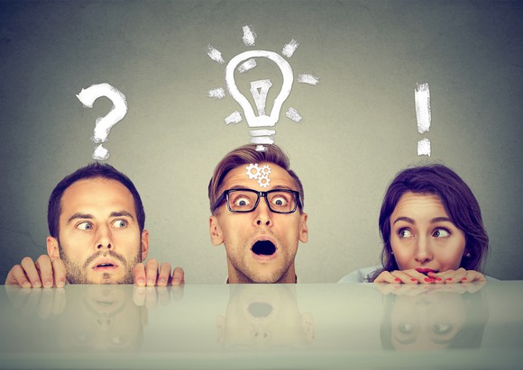 Scared and anxious young people (two men and a woman) who are hiding peek from under the table. Icons above their heads indicate that they have questions and ideas to solve a problem.