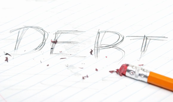 Part of a pencil is shown, after it has started erasing the word debt on a sheet of lined paper