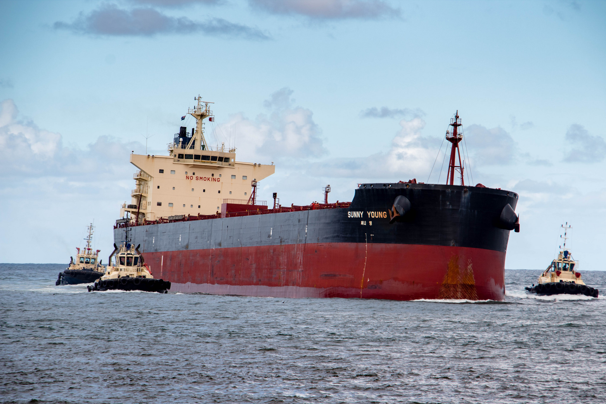 A cargo ship is steered by two small tugboats on the ocean. 