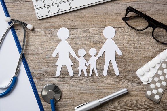 A paper cutout of a family on a desk next to a stethoscope, pills, a pair of glasses, a pen, and a keyboard.