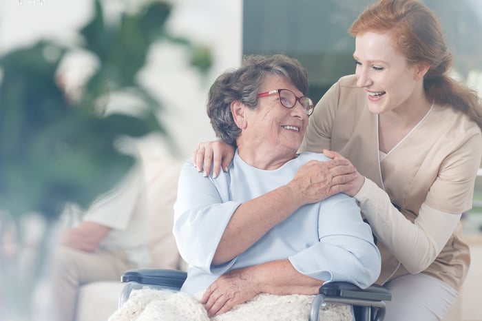 Older woman in wheelchair smiling and holding hands with younger woman in scrubs.